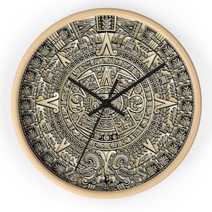 May include: A round wall clock with a light wood frame featuring a detailed, metallic gold and black graphic print of the Aztec Sun Stone calendar. The clock face displays intricate carved patterns, symbols, and faces, with three black hands.