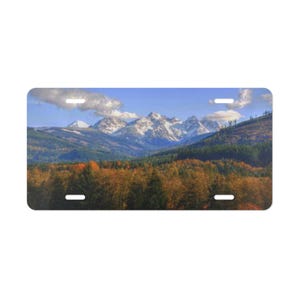 Könnte beinhalten: Ein rechteckiges Nummernschild mit einer Landschaftsaufnahme von schneebedeckten Berggipfeln unter einem klaren blauen Himmel mit vereinzelten weißen Wolken. Im Vordergrund ist ein dichter Wald mit herbstlich gefärbtem Laub zu sehen.