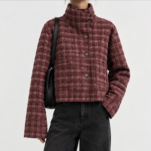 Puede incluir: Una chaqueta corta acolchada con un patrón de cuadros en rojo oscuro y marrón, que presenta un cuello alto, mangas largas y un cierre frontal de botones. La chaqueta se combina con pantalones vaqueros gris oscuro y un bolso negro.