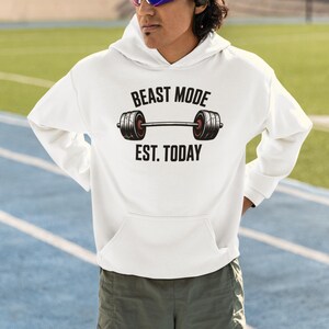 Könnte beinhalten: Ein weißer Kapuzenpullover mit einer Grafik einer schwarzen Langhantel mit roten Akzenten. Der Text über der Hantel lautet BEAST MODE und der Text darunter EST. TODAY. Der Hoodie verfügt über lange Ärmel, eine Kapuze und eine Kängurutasche.