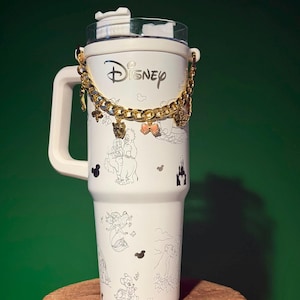 Könnte beinhalten: Ein weißer Isolierbecher mit Henkel und durchsichtigem Deckel, verziert mit schwarzen Strichzeichnungen verschiedener Disney-Figuren und dem Disney-Logo. Eine goldfarbene Metallkette mit kleinen Anhängern ist am Becher befestigt.