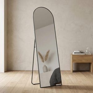 May include: A full-length floor mirror featuring a tall, arched top design with a thin, black metal frame. The mirror is supported by a slim, black metal stand attached to the back, allowing it to stand upright independently on the floor.