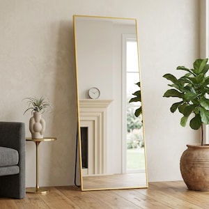 May include: A tall, rectangular floor mirror featuring a thin, metallic gold frame. The mirror is positioned upright, reflecting the interior space, including a fireplace and a clock. The design is simple and functional, with a clean, reflective surface.