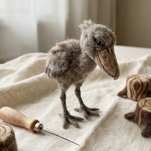 May include: A needle-felted gray shoebill stork figurine with a textured wool finish, dark eyes, and a large beak, accompanied by three small felted tree stump shapes and a metal felting needle with a wooden handle for crafting projects.