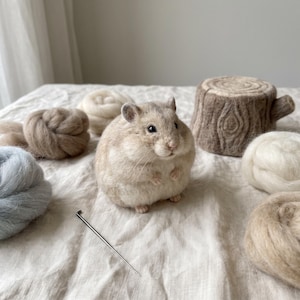 Könnte beinhalten: Eine nadelgefilzte Hamsterfigur in Beige- und Weißtönen, platziert zwischen mehreren Wollvliesknäueln in Weiß, Beige und Hellblau, zusammen mit einem kleinen gefilzten Baumstumpf und einer einzelnen Filznadel aus Metall auf einem strukturierten Stoff.