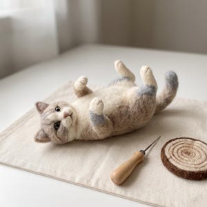 Könnte beinhalten: Eine nadelgefilzte Katzenfigur, die auf dem Rücken liegt und die Pfoten nach oben streckt. Die Katze hat cremefarbenes, hellbraunes und graues Fell, dunkle Augen und eine rosa Nase. Daneben liegen eine Filznadel mit Holzgriff und eine braune Filzscheibe.