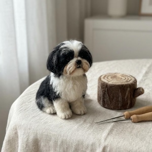 May include: A needle-felted Shih Tzu dog figurine with black and white fur, sitting upright. Beside the dog is a small, cylindrical brown felted tree stump prop and two metal felting needles with wooden handles resting on the table surface.