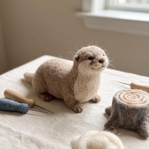 May include: A needle-felted otter figurine with light brown and cream-colored wool fur, featuring a small dark nose and eyes. Beside the otter are two felting needles with wooden handles, a small felted tree stump, and a tuft of white wool.