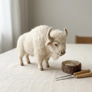 May include: A needle-felted cream-colored bison figurine with a textured wool surface, curved horns, and a small snout. Beside the bison sits a small, round, brown felted log-shaped object and two thin metal needles with light wooden handles.