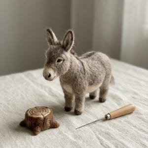 Peut inclure: Une figurine d'âne en laine feutrée à la texture grise et blanche, placée à côté d'une petite souche d'arbre en laine feutrée et d'une aiguille à feutrer en métal avec un manche en bois. L'âne a des oreilles droites et un museau clair.