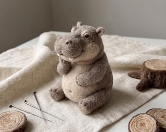 Op de afbeelding: Een naaldgevilte nijlpaardfiguur met een textuur van lichtbruine en beige wol, voorzien van kleine zwarte ogen, twee zichtbare witte tanden en afgeronde oren. Het figuurtje bevindt zich in een zittende houding met kleine ledematen.