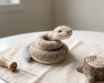 May include: A needle-felted beige and light brown coiled snake figurine with a small light blue tail tip, positioned next to a wooden-handled felting tool and a small, round, brown and beige felted tree stump with circular ring patterns.