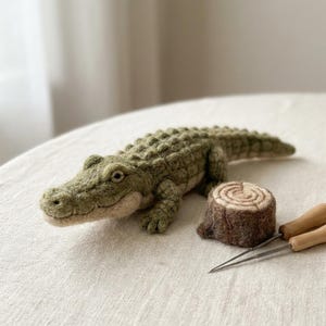 May include: A needle-felted green crocodile figurine with a textured back and a light beige underbelly, positioned next to a small, round, brown felted tree stump and two metal felting needles with wooden handles.