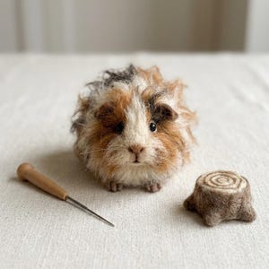 May include: A needle-felted guinea pig figurine with long, textured fur in shades of white, brown, and black. Beside the figurine sits a small, round, brown felted tree stump and a wooden-handled metal felting needle tool.