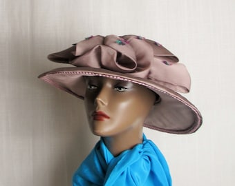 May include: A mauve-colored hat featuring a wide brim and a large, layered fabric flower detail on the crown, accented with small, colorful embroidered floral motifs. The hat is paired with a bright blue scarf draped around the neck area.