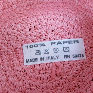 May include: Close-up of a pink woven hat label that reads "100% PAPER MADE IN ITALY RN 59478". The label has care instructions with symbols for hand wash, do not bleach, do not tumble dry, and iron at low temperature.
