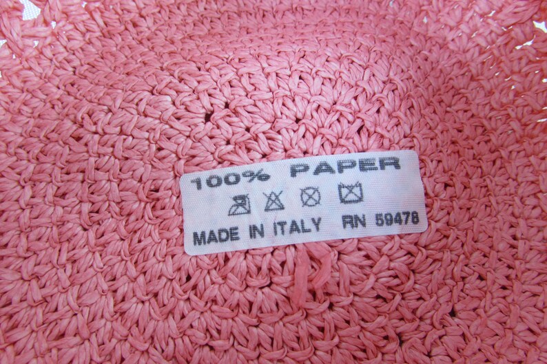 May include: Close-up of a pink woven hat label that reads "100% PAPER MADE IN ITALY RN 59478". The label has care instructions with symbols for hand wash, do not bleach, do not tumble dry, and iron at low temperature.