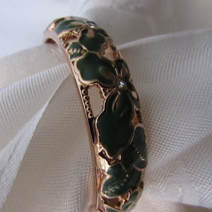 May include: A gold-toned bangle bracelet with a green enamel floral design. The flowers are intricate and detailed, with a small clear rhinestone in the center of each flower.