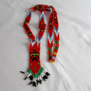 Op de afbeelding: Een lange kralenketting met een rode achtergrond en geometrische patronen in blauw, wit, geel en groen. De centrale hanger toont een zwart vogelmotief en de onderkant is afgewerkt met een franje van witte en zwarte kralen.