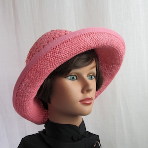 May include: A pink straw hat with a wide brim and a crocheted design. The hat is worn by a mannequin with brown hair.