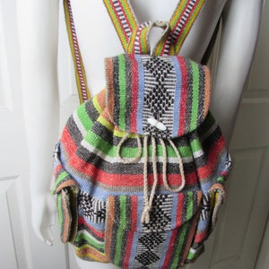 Könnte beinhalten: Ein bunter, gestreifter, gewebter Rucksack mit Kordelzugverschluss und zwei Fronttaschen. Der Rucksack hat einen weißen Knopf und braune Träger.