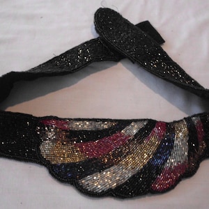 May include: A black beaded belt with a scalloped design. The belt features a central section with colourful beaded stripes in gold, silver, red, and blue. The belt is approximately 7.6 cm wide.