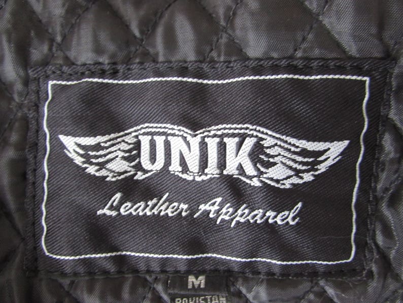 May include: Black leather apparel label with white stitching. The label features a white winged logo with the text "UNIK Leather Apparel" in white.