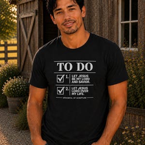 May include: A black short-sleeved t-shirt featuring white text that reads TO DO, 1. LET JESUS BE MY LORD AND SAVIOR, 2. LET JESUS LORD OVER MY LIFE, and SPOONFUL OF SCRIPTURE, with two checkbox icons containing checkmarks next to the numbered items.