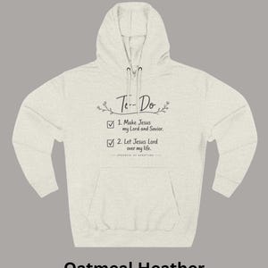 May include: An oatmeal heather hoodie with a front kangaroo pocket and drawstring hood. The chest features black text that reads To-Do, 1. Make Jesus my Lord and Saviour, 2. Let Jesus Lord over my life, and Spoonful of Scripture with floral accents.