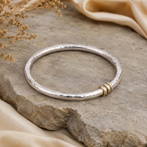 May include: A silver bangle bracelet with a hammered texture featuring three small, smooth gold-toned rings that slide along the band. The bangle has a circular shape and a polished finish, contrasting the silver metal with gold accents.