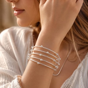 May include: A multi-strand silver bracelet featuring five thin, polished metallic bands. Each band is accented with small, evenly spaced spherical silver beads. The design creates a layered, stacked appearance around the wrist with a bright, reflective finish.