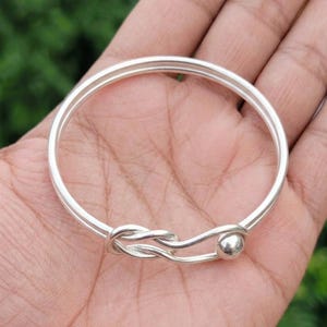 May include: A silver-colored bangle bracelet featuring a double-wire design that converges into a decorative knot detail on one side, finished with a small, smooth, round silver-colored bead at the end of the wire structure.