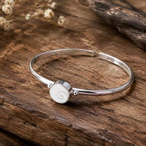May include: A silver cuff bracelet featuring a circular centrepiece with a white and grey spiral pattern. The bracelet has a polished metallic finish and a small bead detail on each side of the central circular element, with an open back design.