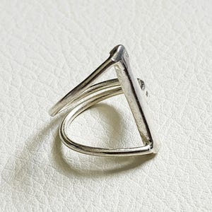 May include: A silver-colored ring featuring a geometric triangular shape. The band is constructed from a single continuous piece of metal that forms an open, angular frame, creating a distinct three-sided silhouette with a polished metallic finish.