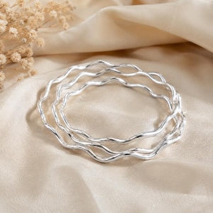 May include: A set of three silver-toned bangles, each featuring a continuous wavy or rippled design. The bracelets are circular in shape with a polished, reflective metallic finish that catches the light, creating a smooth and uniform appearance.