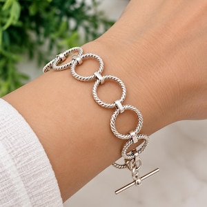 May include: A silver-toned bracelet composed of a series of interconnected circular links with a twisted rope-like texture. The bracelet features a toggle clasp closure consisting of a straight metal bar and a circular ring at the end.