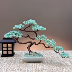 May include: A decorative bonsai tree sculpture crafted from twisted dark copper wire and small, light green gemstone chips. The tree is set in a white rectangular ceramic pot filled with matching green stones, positioned next to a black lantern.