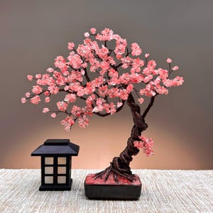May include: A decorative wire bonsai tree featuring numerous small, translucent pink crystal chips as leaves, set in a rectangular black base. Beside the tree stands a small, black, square-shaped lantern with white panels and a grid-like frame.