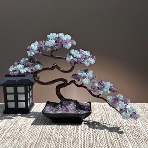 May include: A decorative wire bonsai tree sculpture featuring clusters of light blue and purple gemstone chips as leaves. The tree is anchored in a black rectangular pot filled with purple stones, positioned next to a black lantern-style lamp.