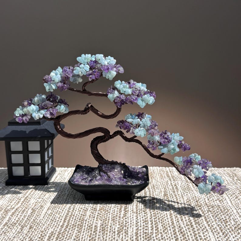 May include: A decorative wire bonsai tree sculpture featuring clusters of light blue and purple gemstone chips as leaves. The tree is anchored in a black rectangular pot filled with purple stones, positioned next to a black lantern-style lamp.