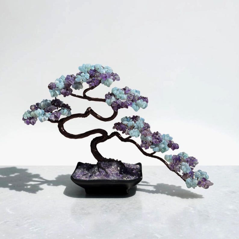 May include: A decorative bonsai tree sculpture featuring twisted dark brown wire branches adorned with clusters of light blue and purple gemstone chips. The tree is anchored in a square black ceramic pot filled with small purple amethyst stones.