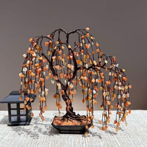 May include: A decorative wire tree sculpture featuring numerous small, tumbled orange and yellow gemstone chips as leaves. The tree has a twisted dark brown wire trunk and branches, set in a square black base filled with matching stone chips.