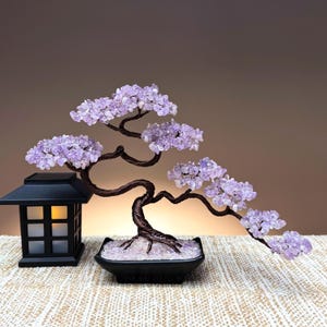May include: A decorative tree sculpture featuring twisted dark brown wire branches adorned with small, light purple amethyst crystal chips. The tree is set in a black rectangular ceramic pot filled with clear crystals, next to a black lantern.