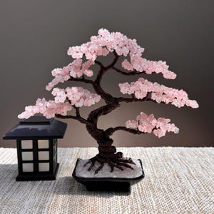 May include: A decorative bonsai tree sculpture featuring twisted dark brown wire branches adorned with small, light pink rose quartz gemstone chips. The tree is planted in a black rectangular ceramic pot filled with white stones, next to a black lantern.