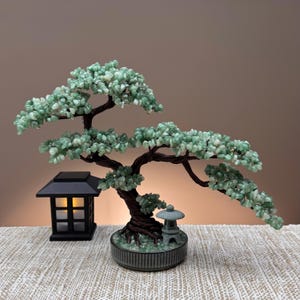 May include: A decorative bonsai tree sculpture featuring twisted dark brown wire branches adorned with small, light green tumbled stone leaves. The tree is set in a round gray pot with a small stone lantern and a black square lantern nearby.