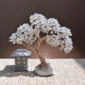 May include: A decorative wire tree sculpture featuring a twisted copper-colored trunk and branches adorned with numerous small, translucent, iridescent white stone chips. The tree is set on a round white base next to a small gray lantern.