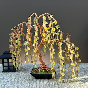 May include: A wire-wrapped sculpture of a weeping willow tree featuring a twisted copper-colored trunk and branches adorned with numerous small, translucent light green gemstone chips. The tree is set in a black rectangular base with green moss.