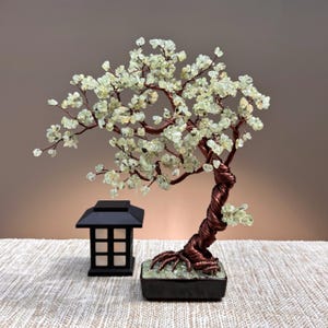 May include: A decorative wire tree sculpture featuring a twisted copper-colored trunk and branches adorned with small, pale green gemstone chips. The tree is rooted in a rectangular black base filled with green stones, next to a small black lantern.