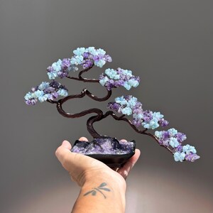 May include: A decorative wire bonsai tree sculpture featuring twisted dark brown metal branches adorned with small, tumbled light blue and purple gemstone chips. The base is a black ceramic dish filled with purple gemstone fragments.