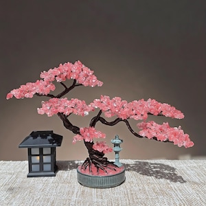 May include: A decorative bonsai tree sculpture featuring twisted dark brown wire branches adorned with small pink crystal leaves, set in a round gray base, accompanied by a small black lantern and a miniature gray stone-style pagoda statue.